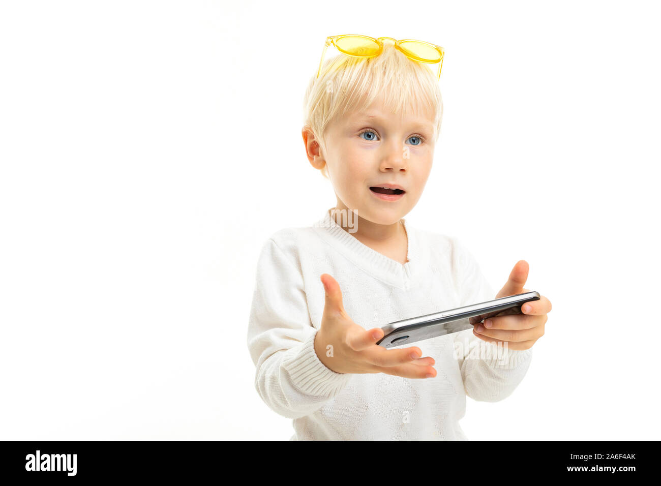 Little boy with short blond hair, blue eyes, cute appearance, in white jacket, light blue pants, stands with yellow glasses and looks into the phone Stock Photo