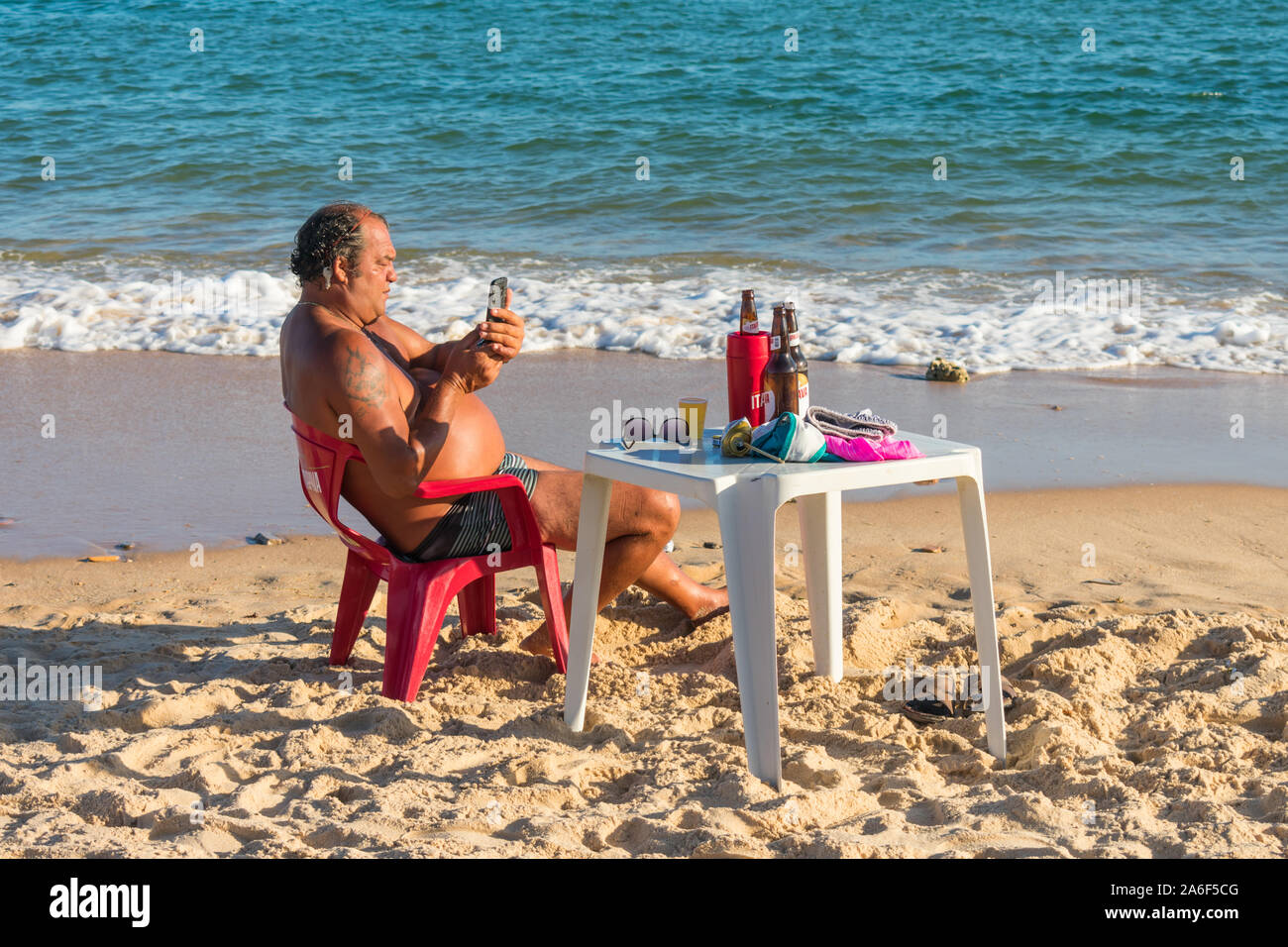 Older man sitting at a bar table by the ocean using his smartphone in Arembepe, Brazil Stock Photo