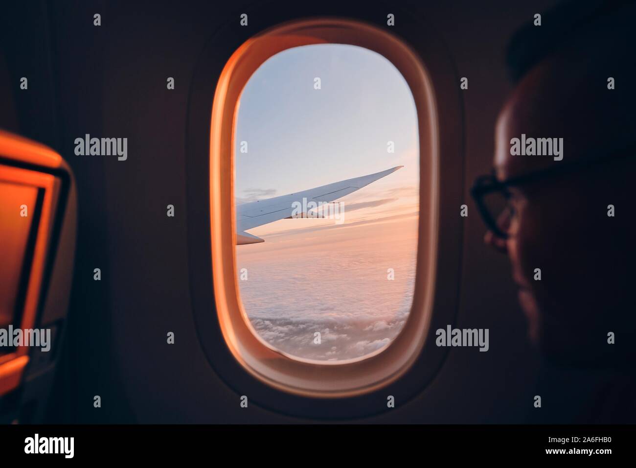 Man looking throught vindow of airplane during flight above clouds at sunset. Selective focus on wing. Stock Photo