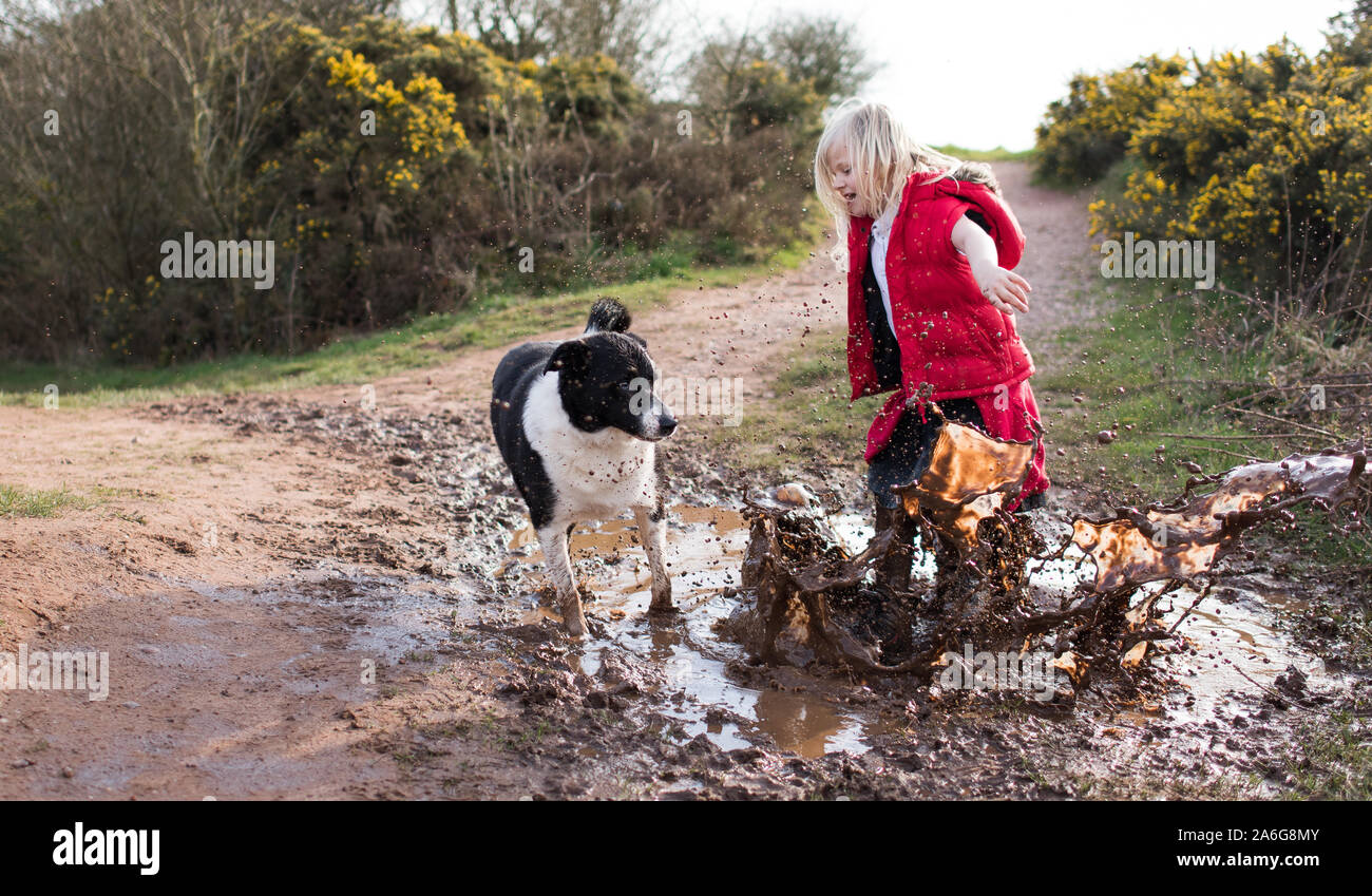 Pretty little girl jumping in very muddy puddle playing with her pet ...