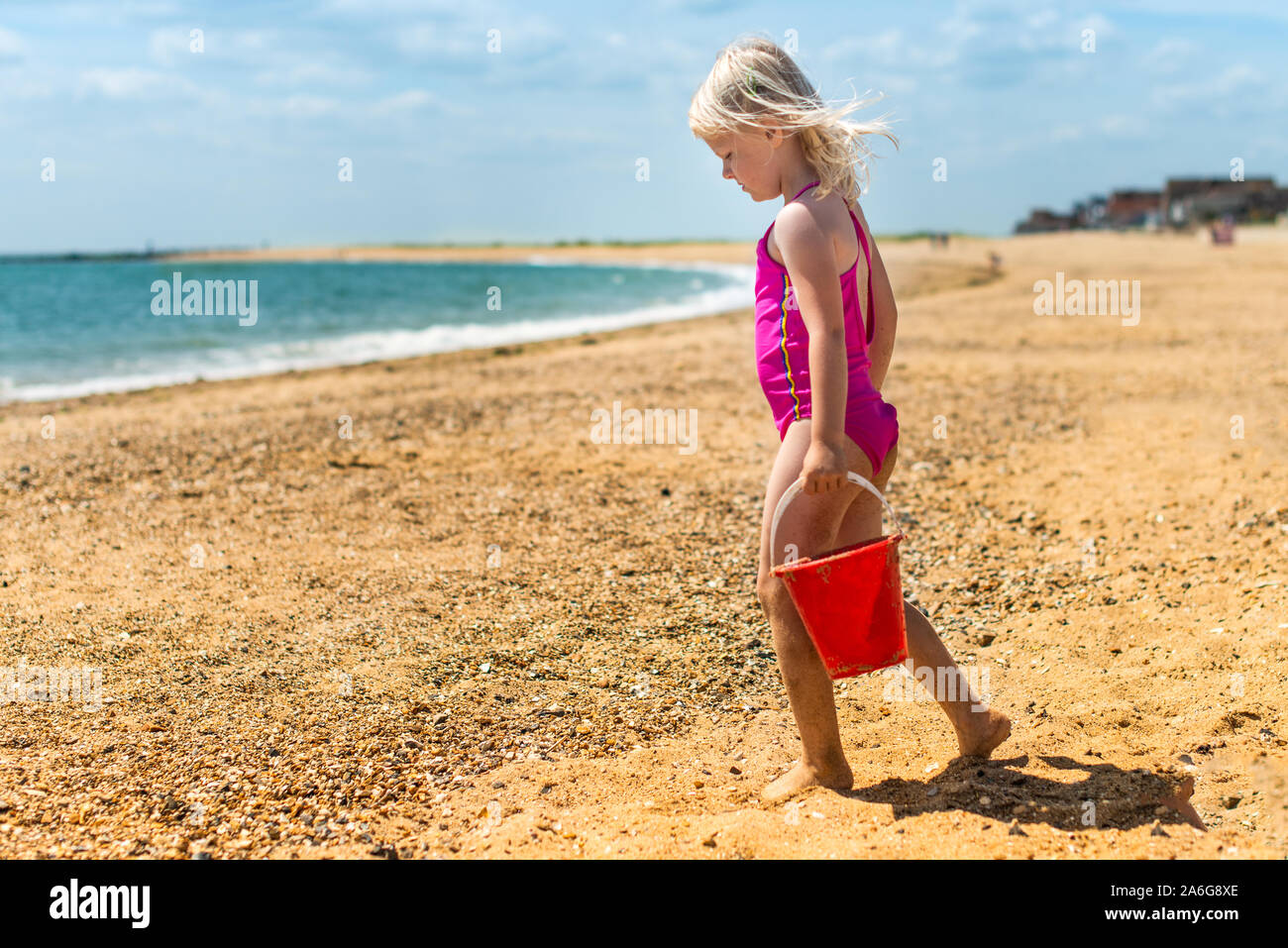 A cute, pretty little girl carrying a red bucket walking on the beach
