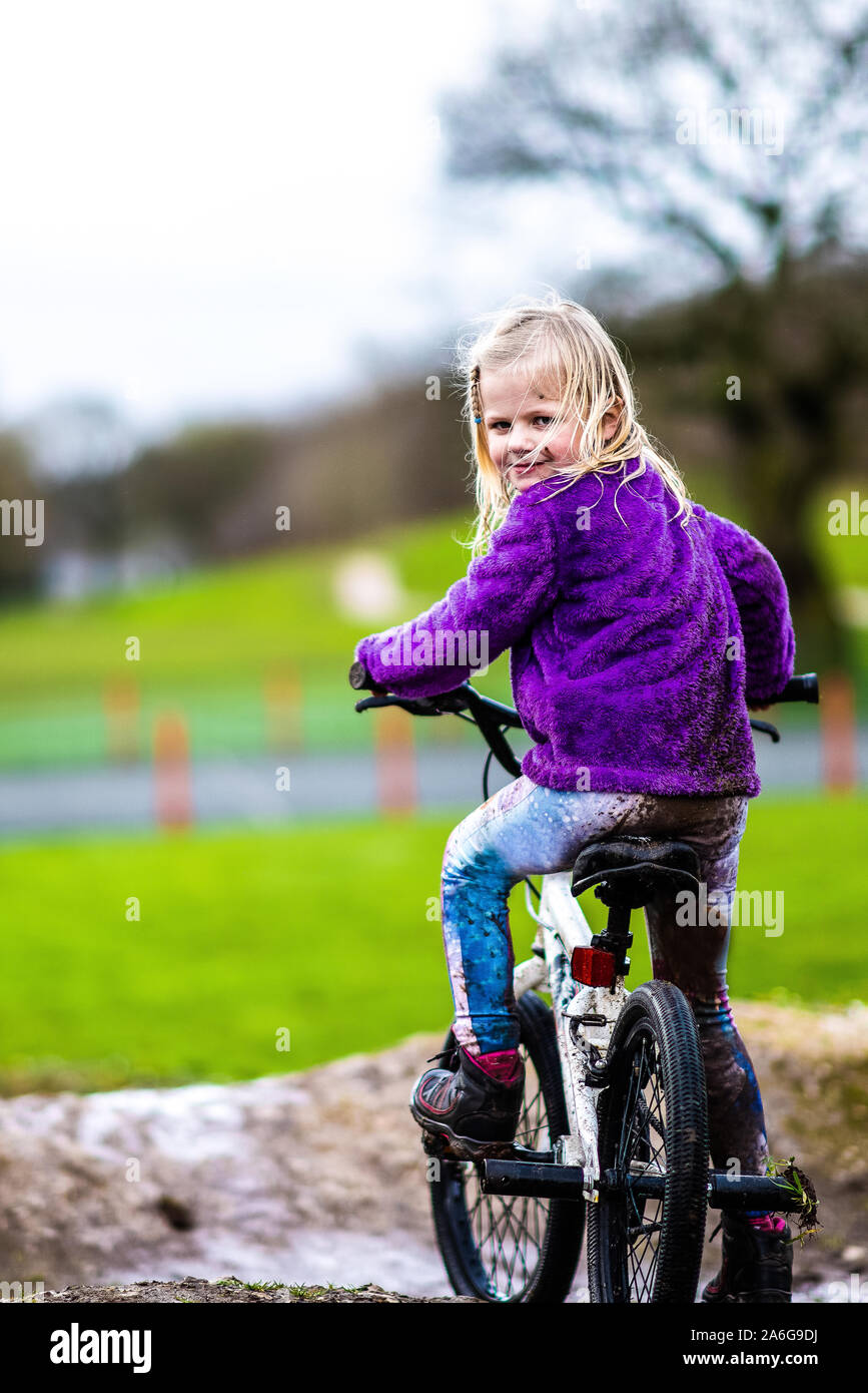 A pretty little girl with blonde hair and a purple jumper enjoys a day at a BMX track, riding and practicing tricks, child in the countryside UK Stock Photo