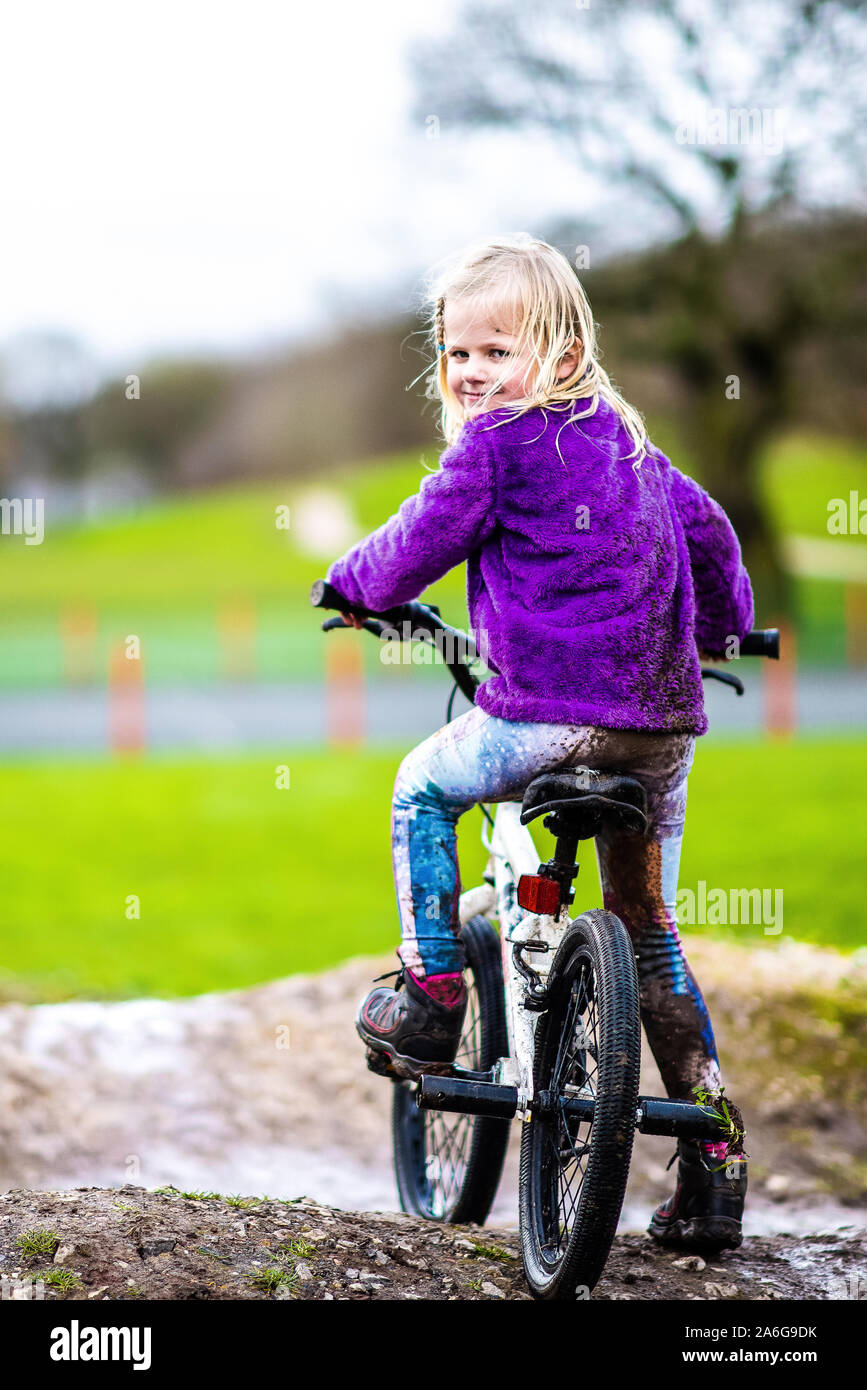 A pretty little girl with blonde hair and a purple jumper enjoys a day at a BMX track, riding and practicing tricks, child in the countryside UK Stock Photo