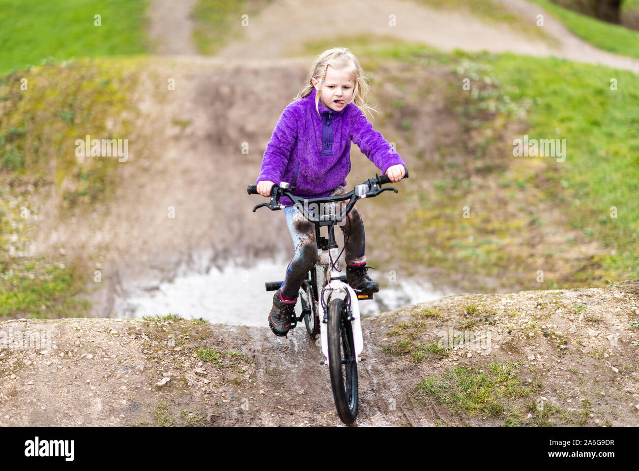 A pretty little girl with blonde hair and a purple jumper enjoys a day at a BMX track, riding and practicing tricks, child in the countryside UK Stock Photo