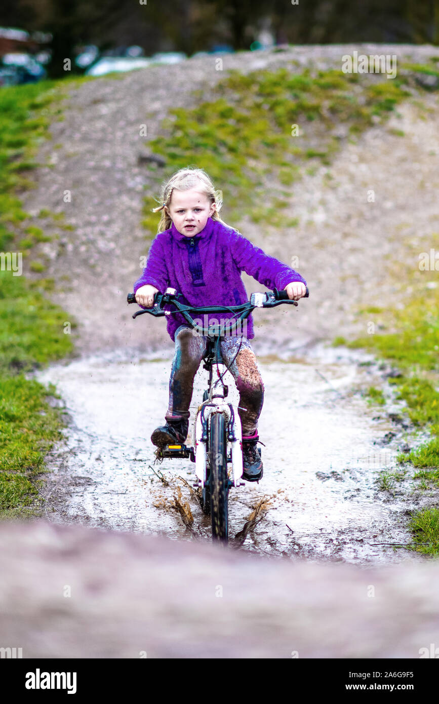 A pretty little girl with blonde hair and a purple jumper enjoys a day at a BMX track, riding and practicing tricks, child in the countryside UK Stock Photo