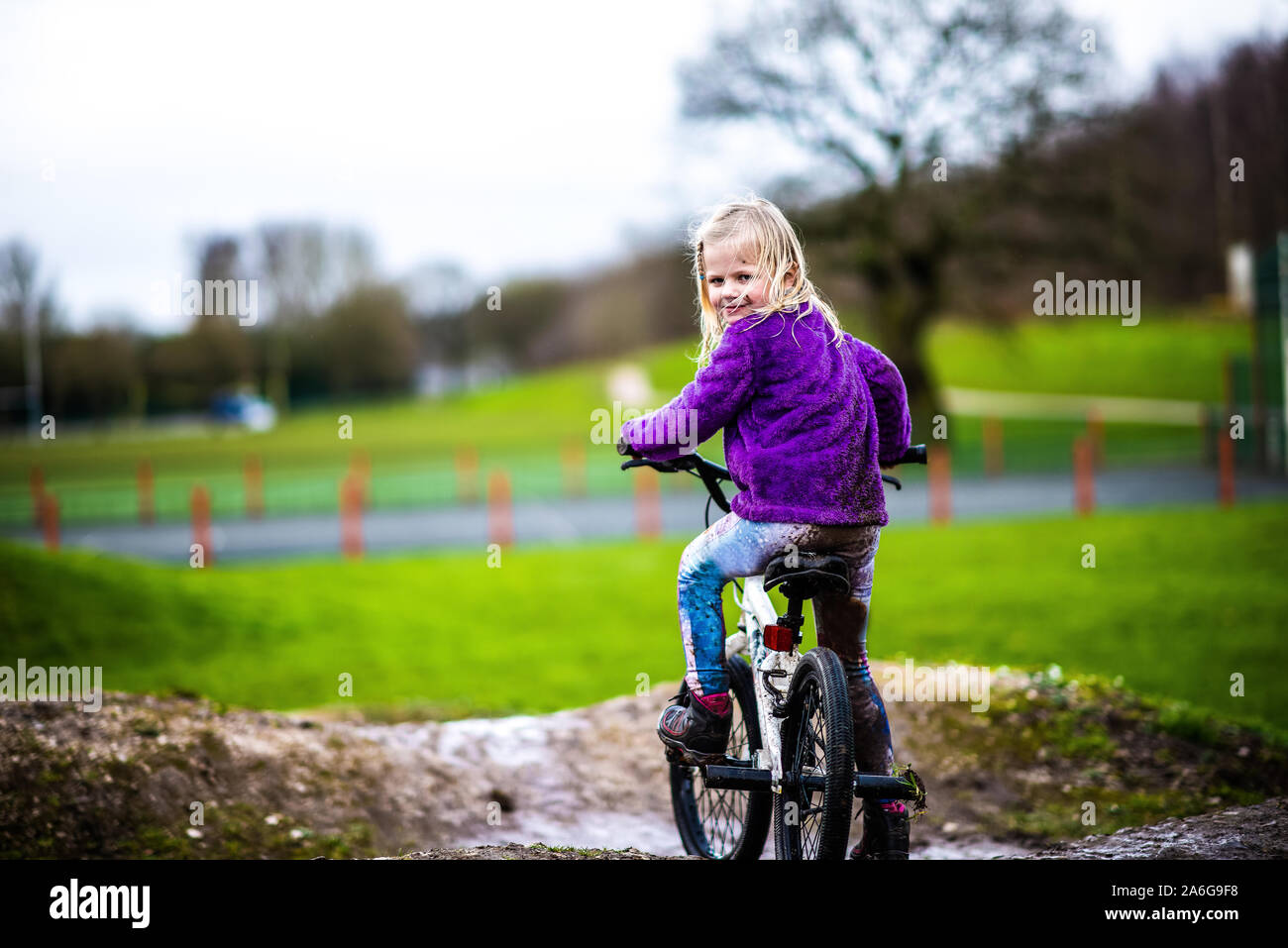 A pretty little girl with blonde hair and a purple jumper enjoys a day at a BMX track, riding and practicing tricks, child in the countryside UK Stock Photo