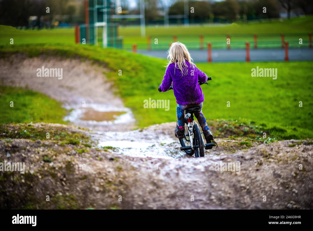 A pretty little girl with blonde hair and a purple jumper enjoys a day at a BMX track, riding and practicing tricks, child in the countryside UK Stock Photo
