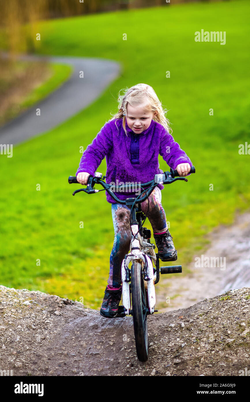 A pretty little girl with blonde hair and a purple jumper enjoys a day at a BMX track, riding and practicing tricks, child in the countryside UK Stock Photo