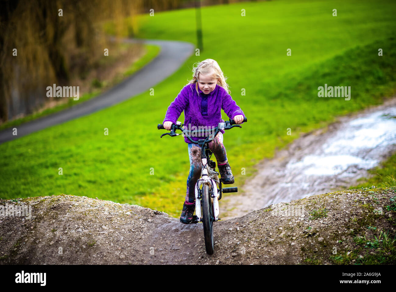 A pretty little girl with blonde hair and a purple jumper enjoys a day at a BMX track, riding and practicing tricks, child in the countryside UK Stock Photo