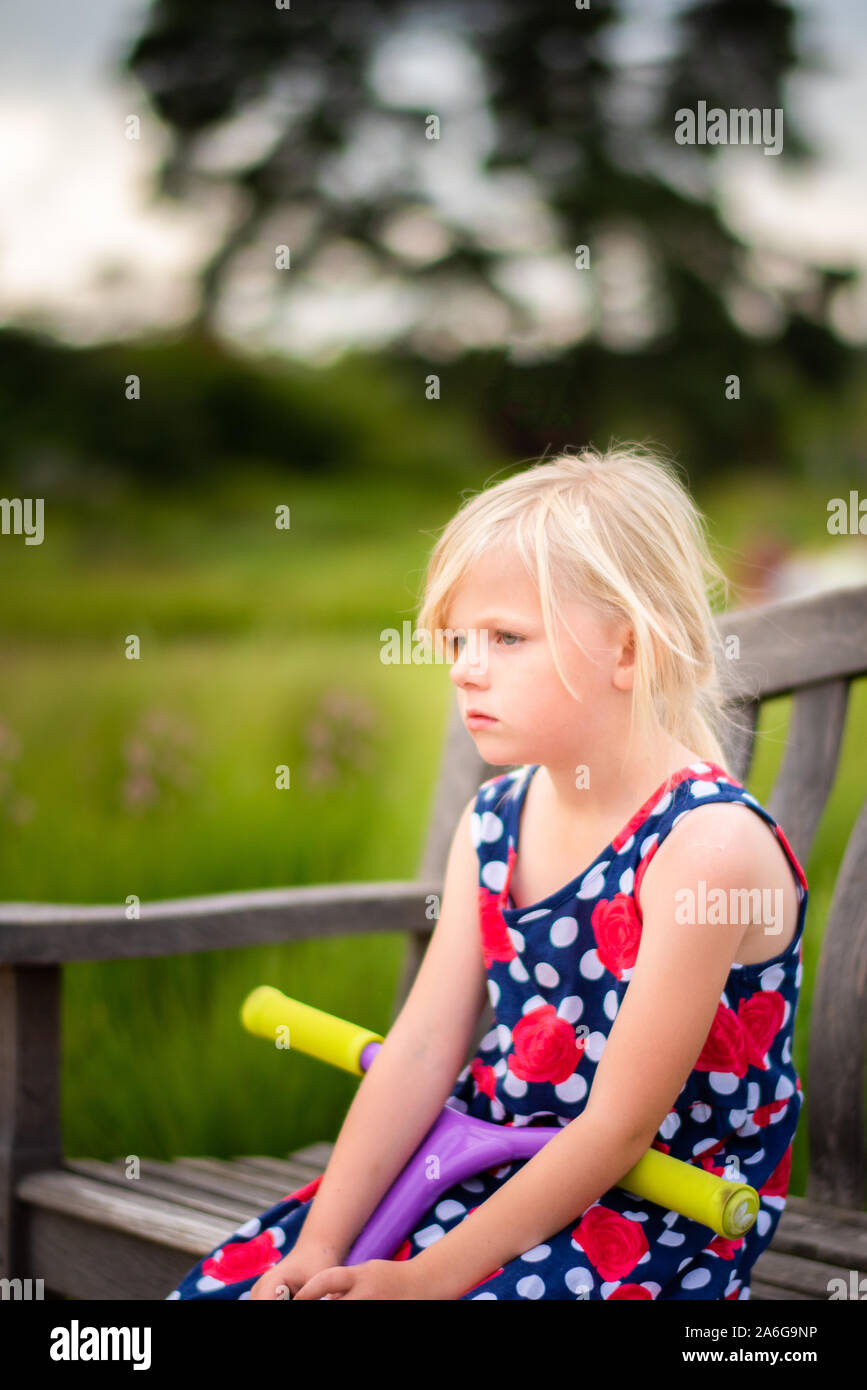 A pretty little girl sitting on a bench wearing a beautiful dress looking and feeling sad after being bullied at School Stock Photo