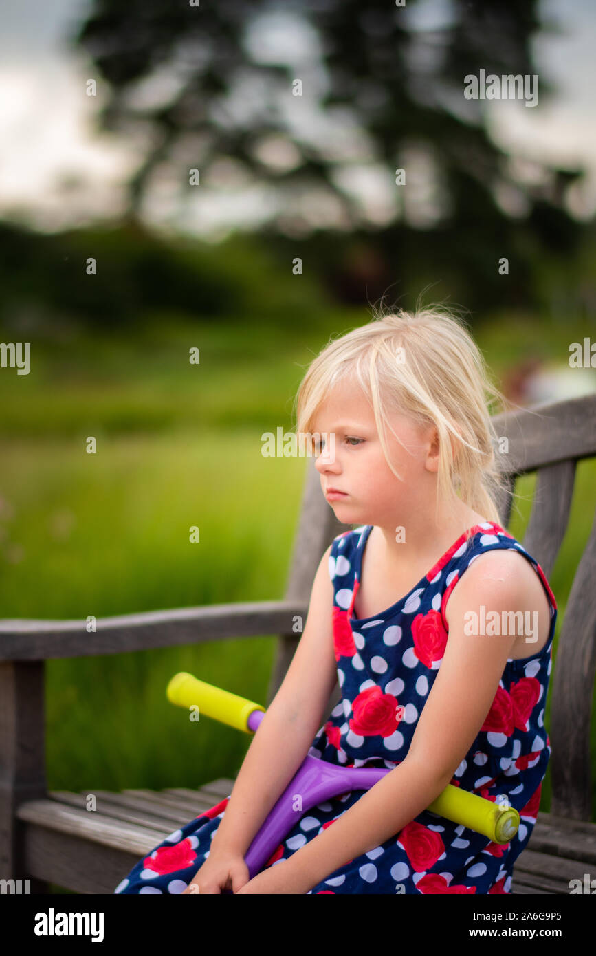 A pretty little girl sitting on a bench wearing a beautiful dress looking and feeling sad after being bullied at School Stock Photo
