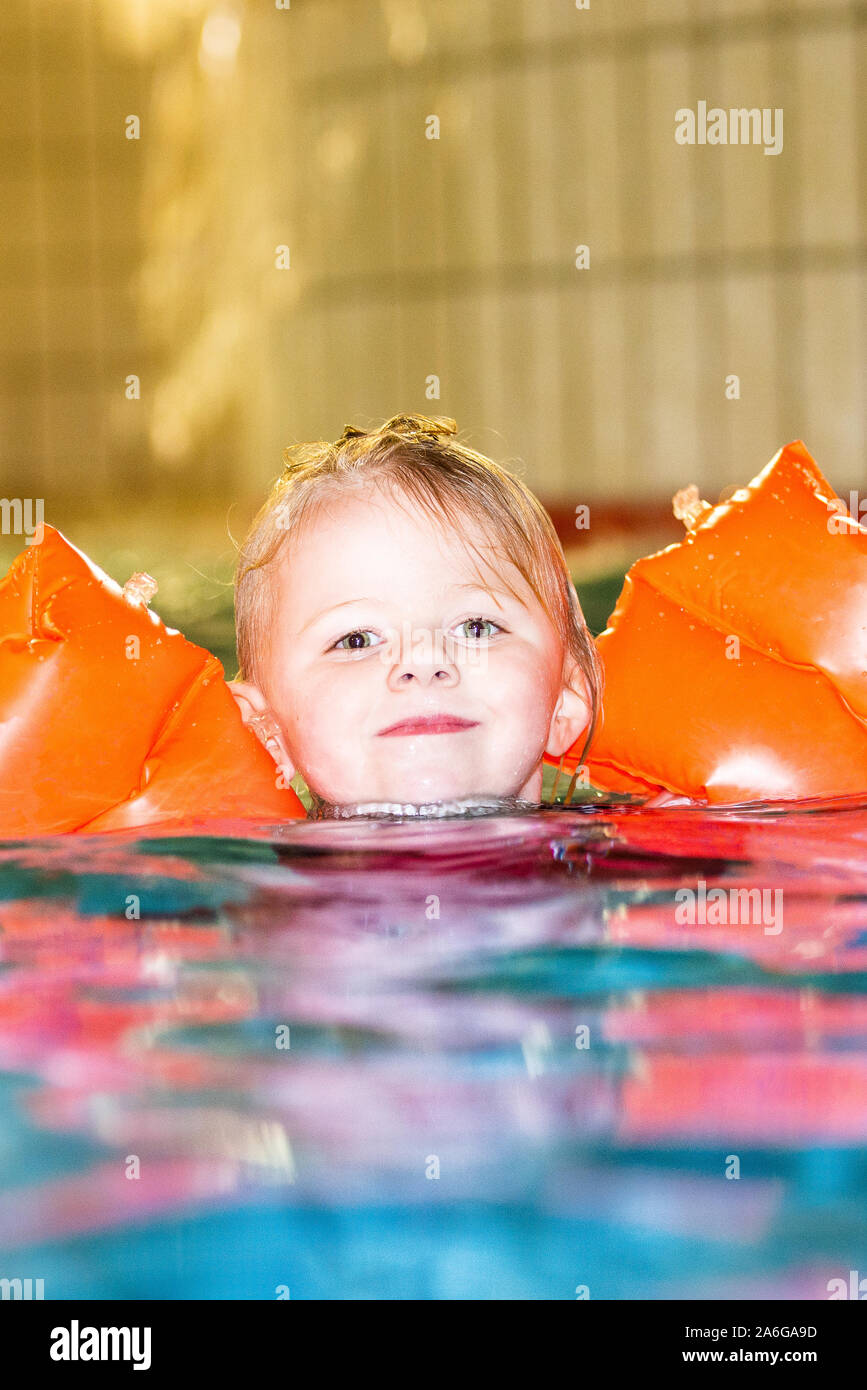 A pretty little girl with orange arm bands smiling and playing happily in the swimming pool, wearing a red swimming costume Stock Photo