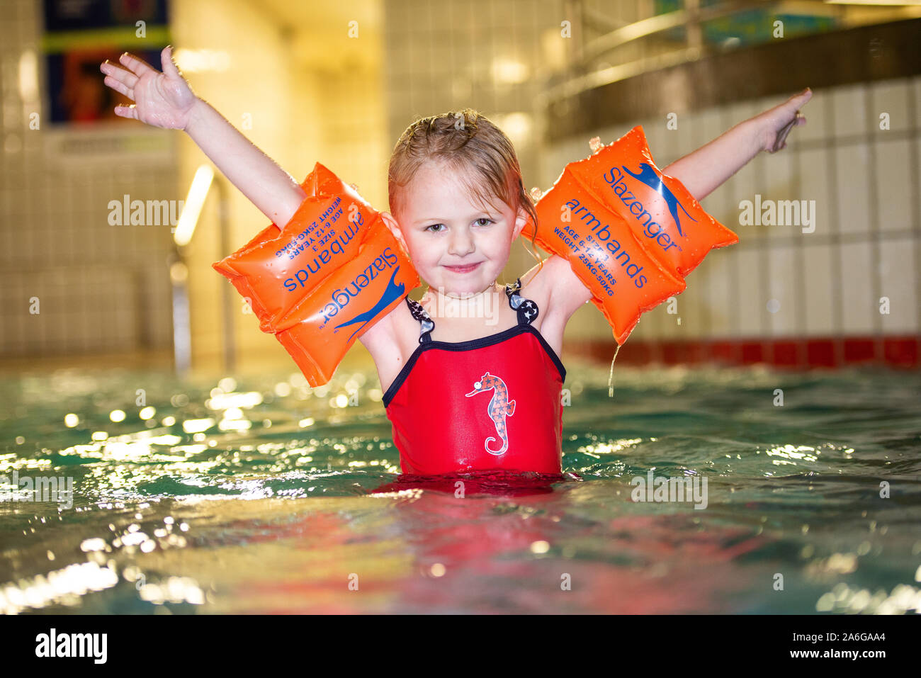 A pretty little girl with orange arm bands and playing happily in the swimming pool, wearing a red swimming costume, Slazenger armbands Stock Photo - Alamy