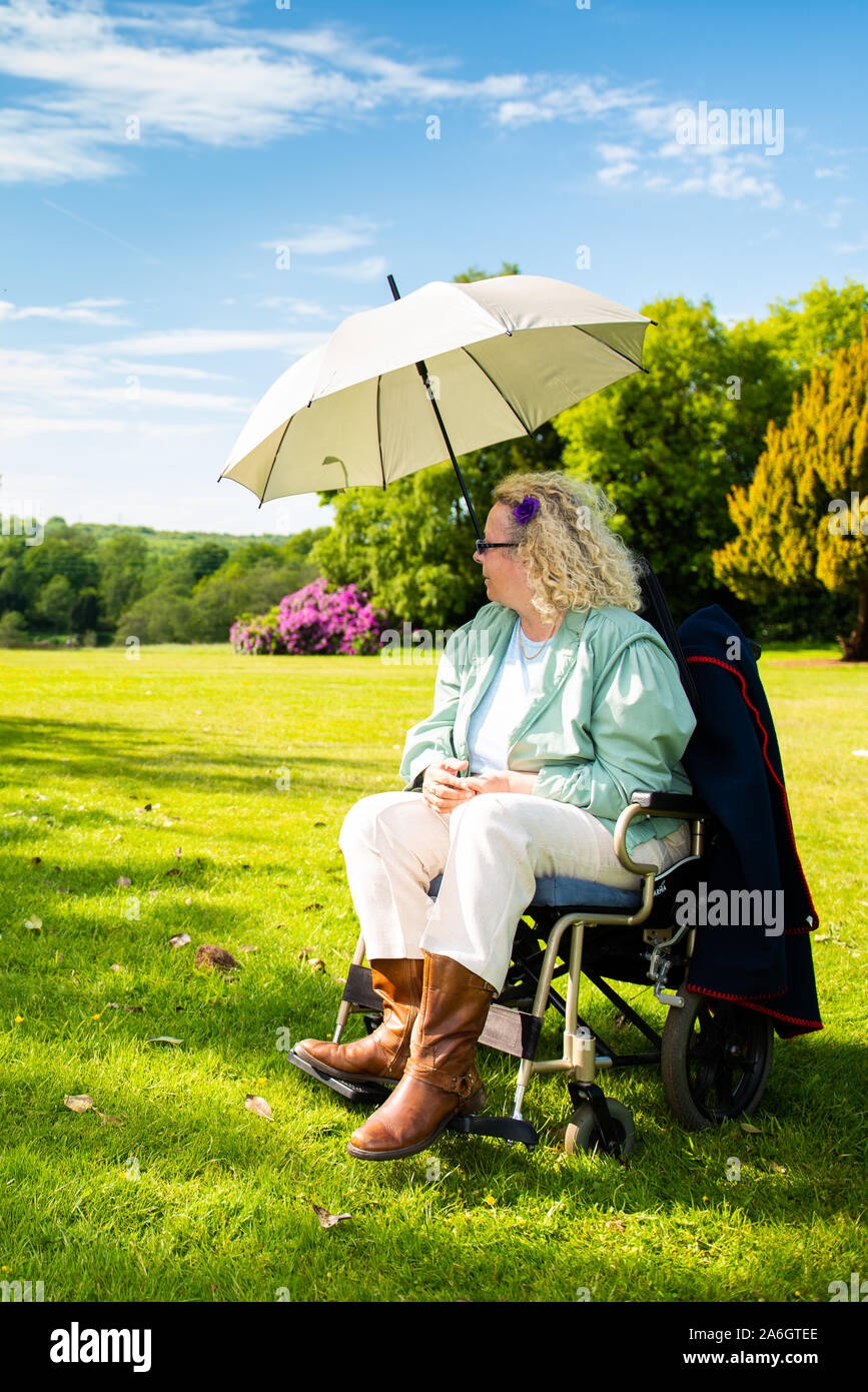 A disabled lady sits in her wheelchair on a hot summers day under an umbrella to keep her cool and in the shade, Stroke, heart attack, illness Stock Photo