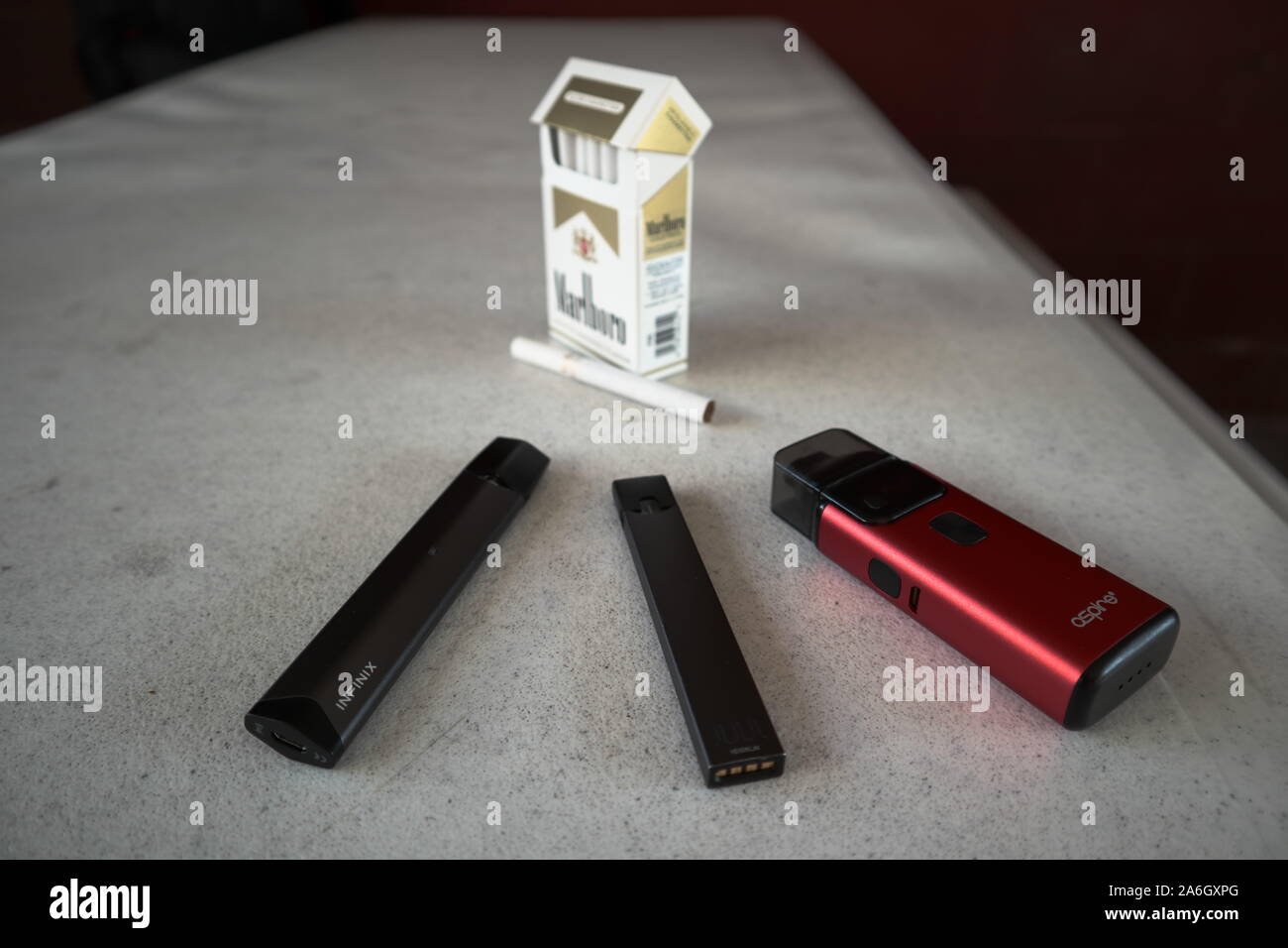 3 vapes juul, aspire breeze, smok infinix with a pack of marlboro cigarettes and one cigarette placed outside on a white textured table, isolated Stock Photo
