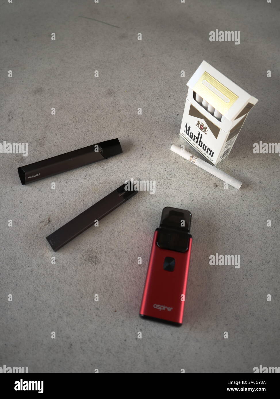 3 vapes juul, aspire breeze, smok infinix with a pack of marlboro cigarettes and one cigarette placed outside on a white textured table, isolated Stock Photo