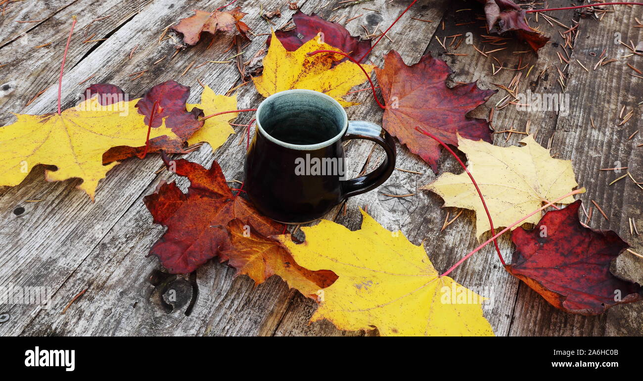 a black mug on a wooden table with colourful fall leaves scattered about.  imperfection and rustic Stock Photo