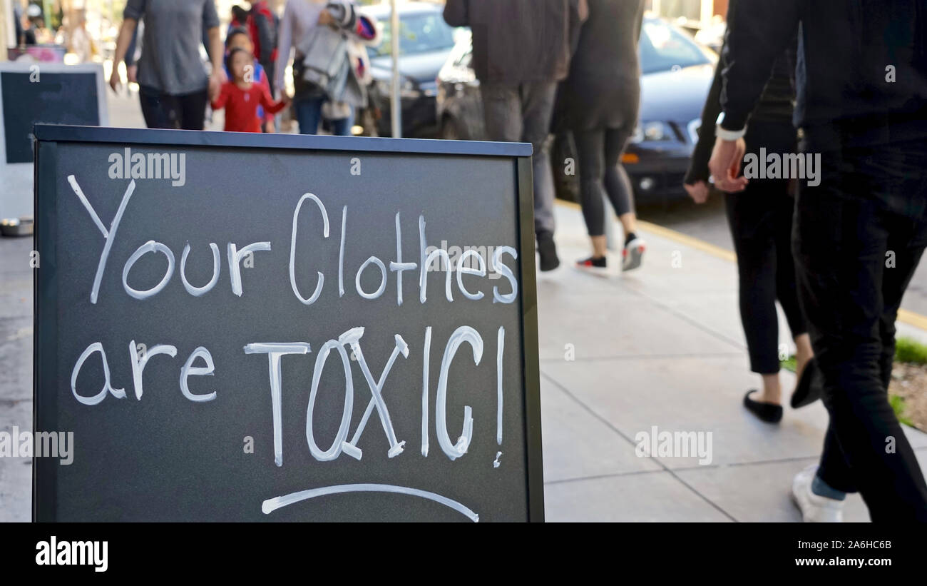 urban sidewalk crowded with pedestrians, foreground hand-lettered black chalkboard sign saying 'Your Clothes Are Toxic!' referring to fast fashion Stock Photo