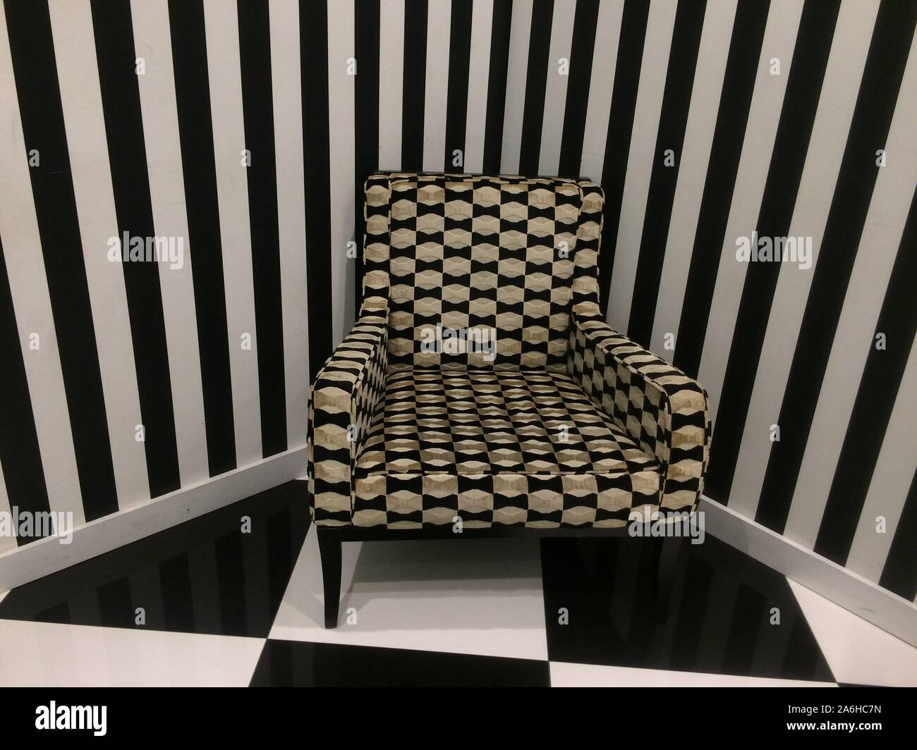 Eccentric modern interior design, monochrome  room with an upholstered corner chair, striped wallpaper and checkboard floor Stock Photo