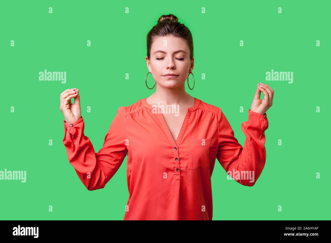 Portrait of peaceful young woman with bun hairstyle, big earrings and in red blouse relaxing and doing meditation gesture with fingers, practicing yog Stock Photo