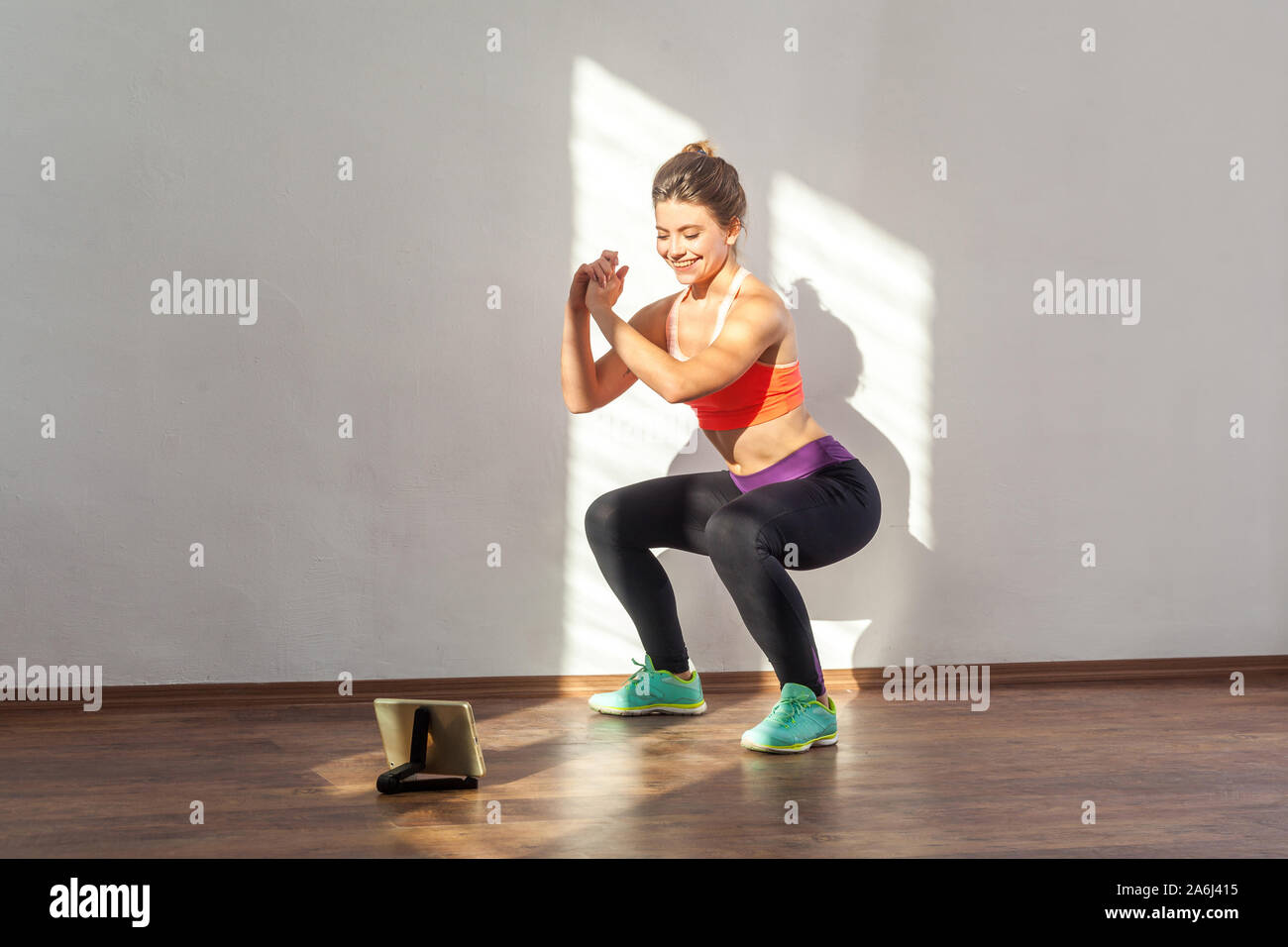 Positive sportive woman with bun hairstyle and in tight sportswear doing squatting sit-up exercise while watching training video on tablet. indoor stu Stock Photo