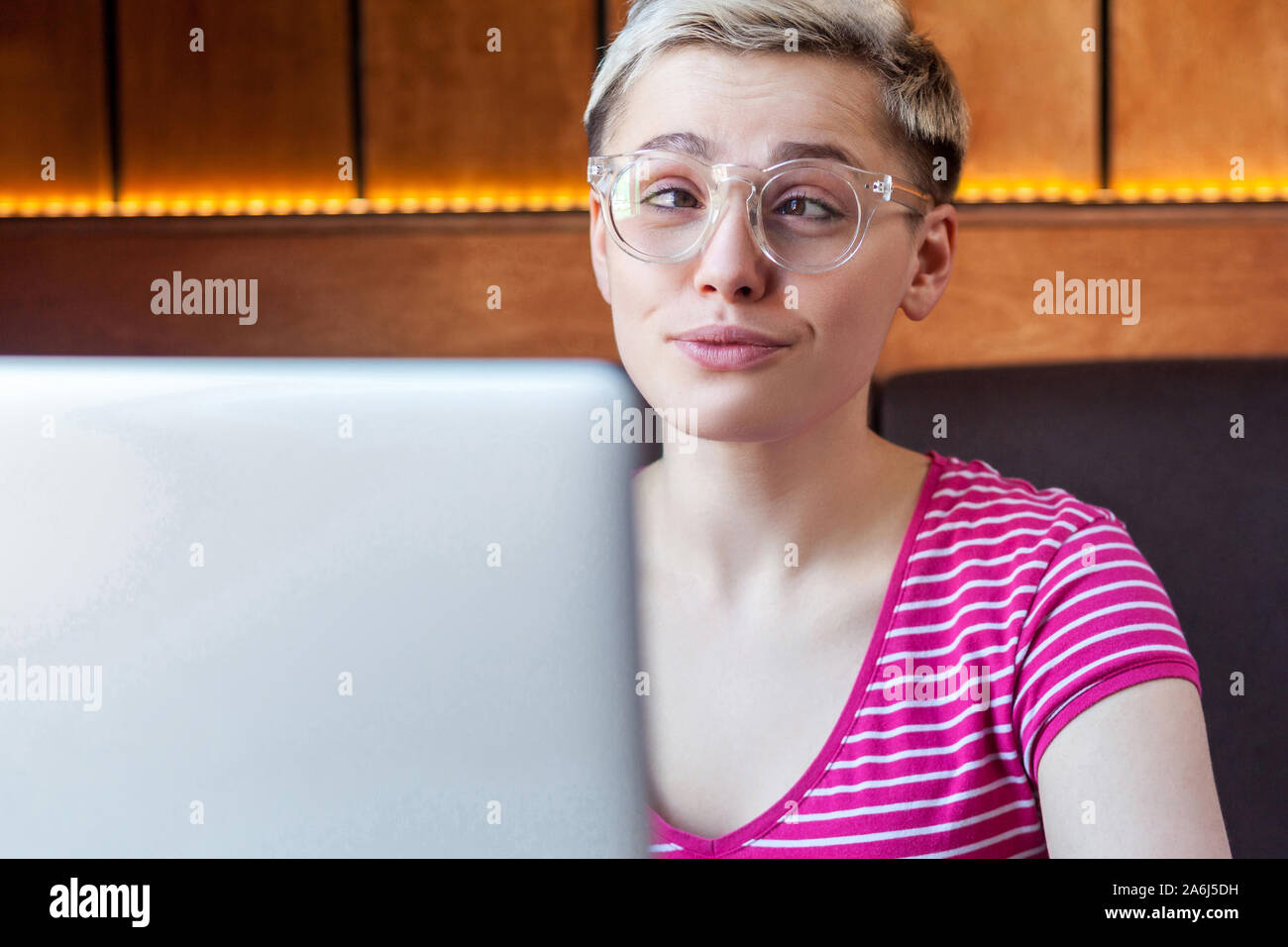 Closeup portrait of happy beautiful childish young girl with short hair in pink t-shirt and eyeglasses is sitting in cafe and working on laptop, cross Stock Photo