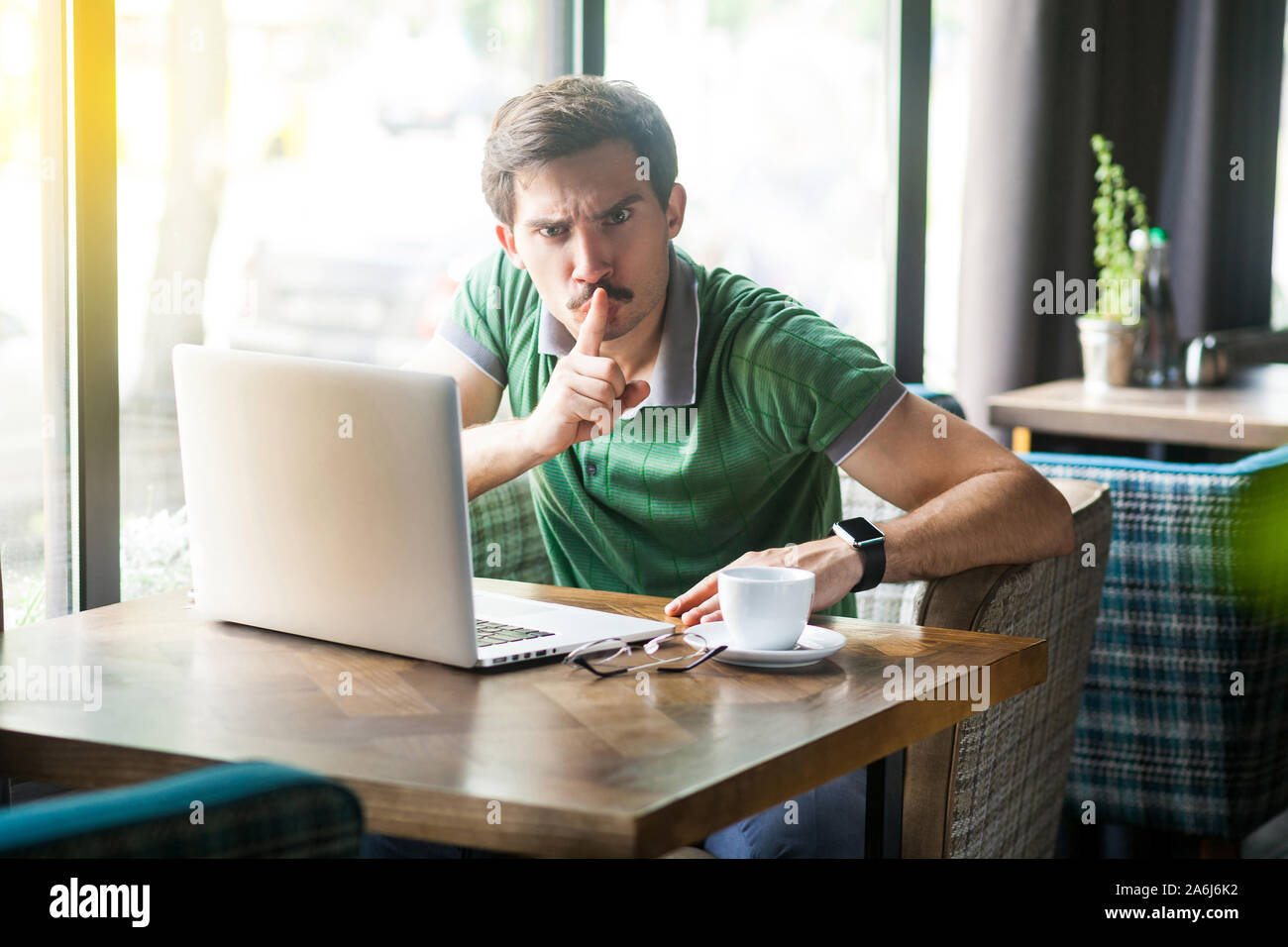 Shhh, Be quiet! Young angry businessman in green t-shirt sitting and working on laptop, looking at camera with finger on lips and command to be silent Stock Photo