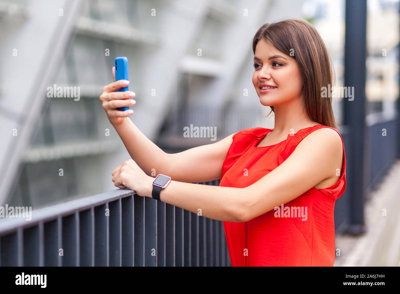 I am beautiful. Half turned portrait of attractive brunette woman in stylish red shirt doing selfie using smartphone, confident in her beauty looking Stock Photo