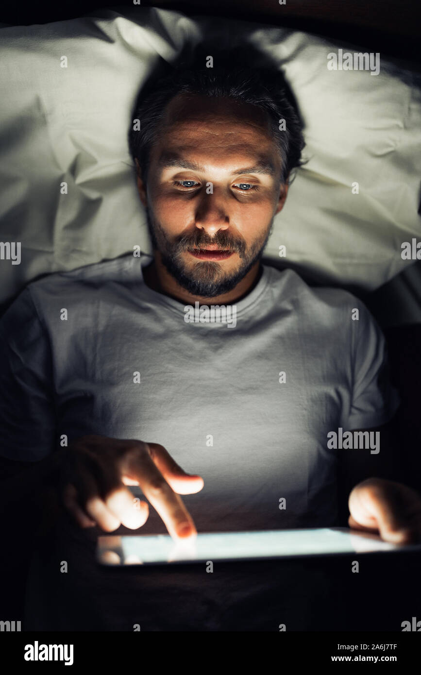 Young handsome and tired man with a beard cannot sleep and is watching something on his tablet at night. Stock Photo