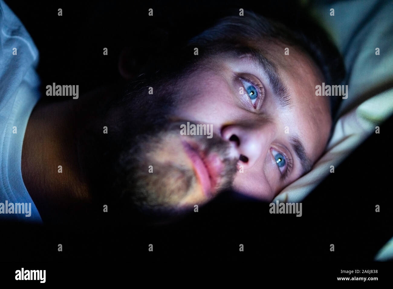 Bearded caucasian male is watching something on his mobile phone in bed at night. He is clearly very tired but cannot sleep. Stock Photo