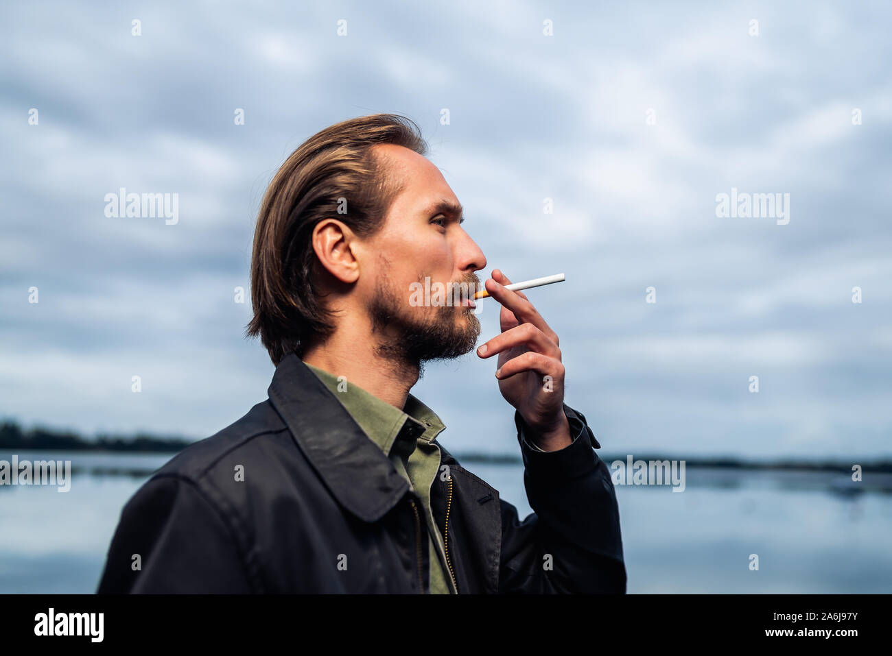 Photo of a young bearded man smoking a cigarette. Lake and clouds in the background. Stock Photo