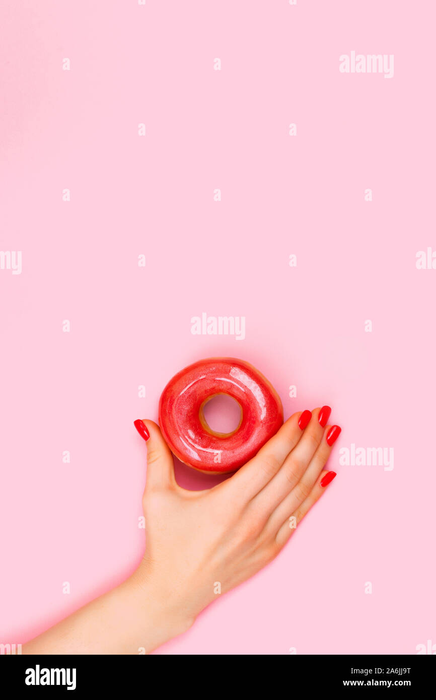 Hand of a beautiful woman holds a sweet red donut with sparkles. Closeup of hands of a young woman with manicure on nails. Stock Photo