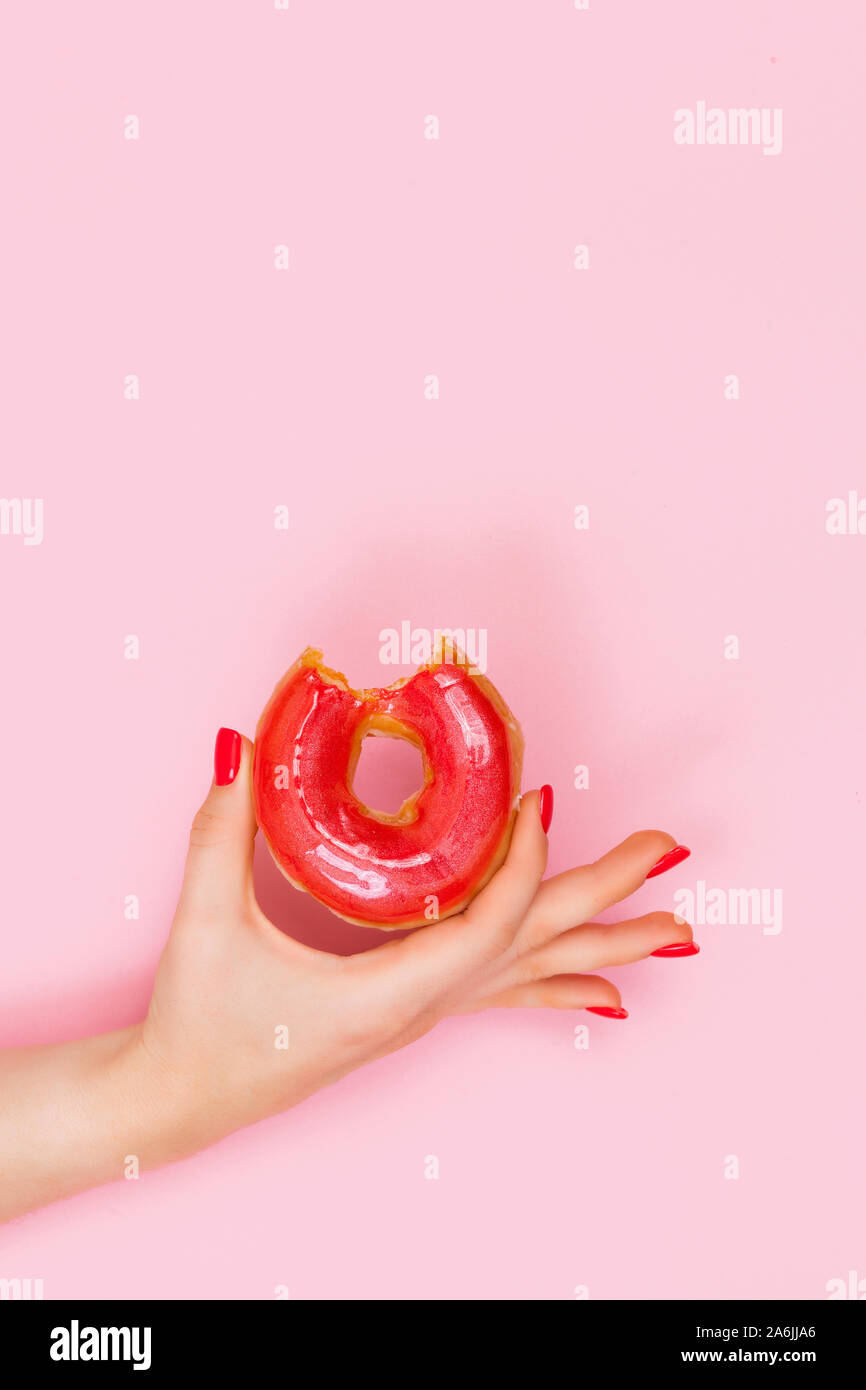 Hand of a beautiful woman holds a sweet red bitten donut with sparkles. Closeup of hands of a young woman with manicure on nails. Stock Photo