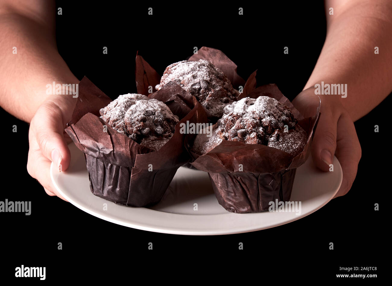 Still life with chocolate muffin with sugar. Woman hand serving or holding a white porcelain plate. Isolated on black background and space for text. Stock Photo