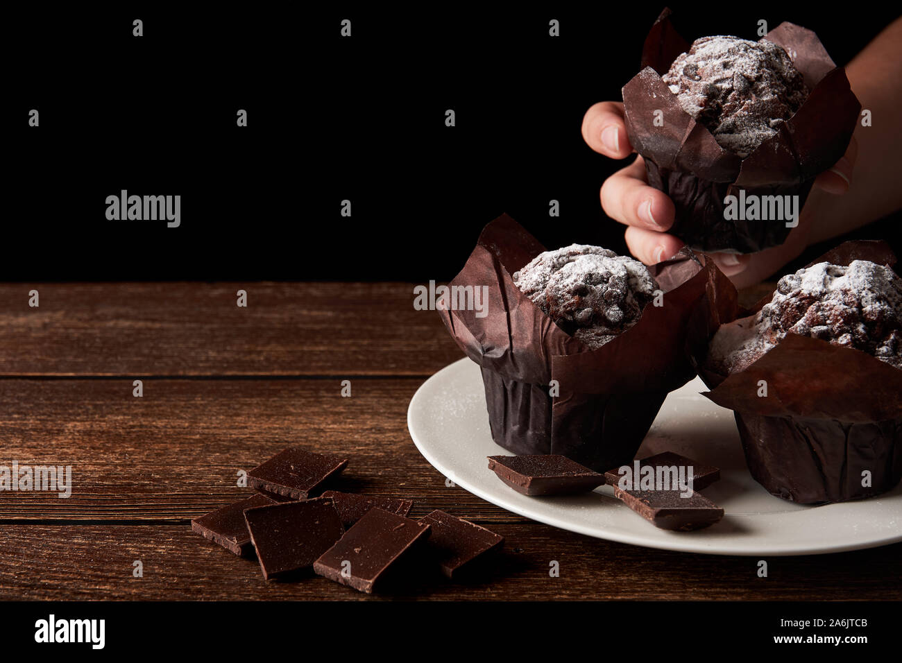 Still life with white porcelain plate and chocolate muffin with sugar on old wooden table top. Woman hand serving or take one dessert. With black back Stock Photo