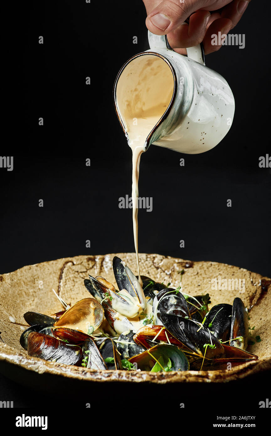 Mussels in creamy white sauce at black background. Chefs hand pours mussels from a jug. Seafood dish Stock Photo