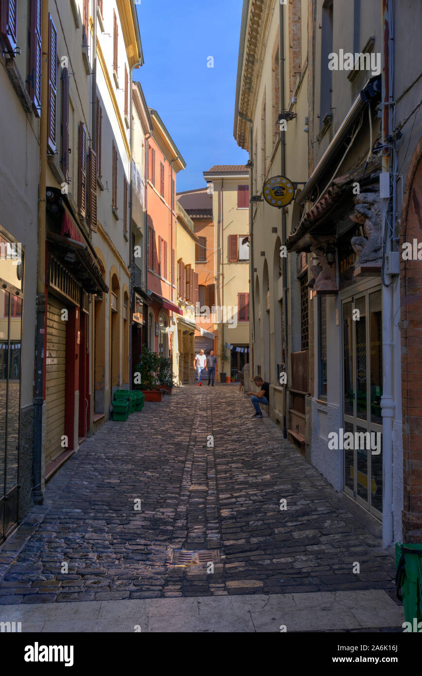 Rimini, Italy - October 20, 2019: Narrow street with cobble stones, two men walking and talking with one man sitting on a step looking at his phone Stock Photo