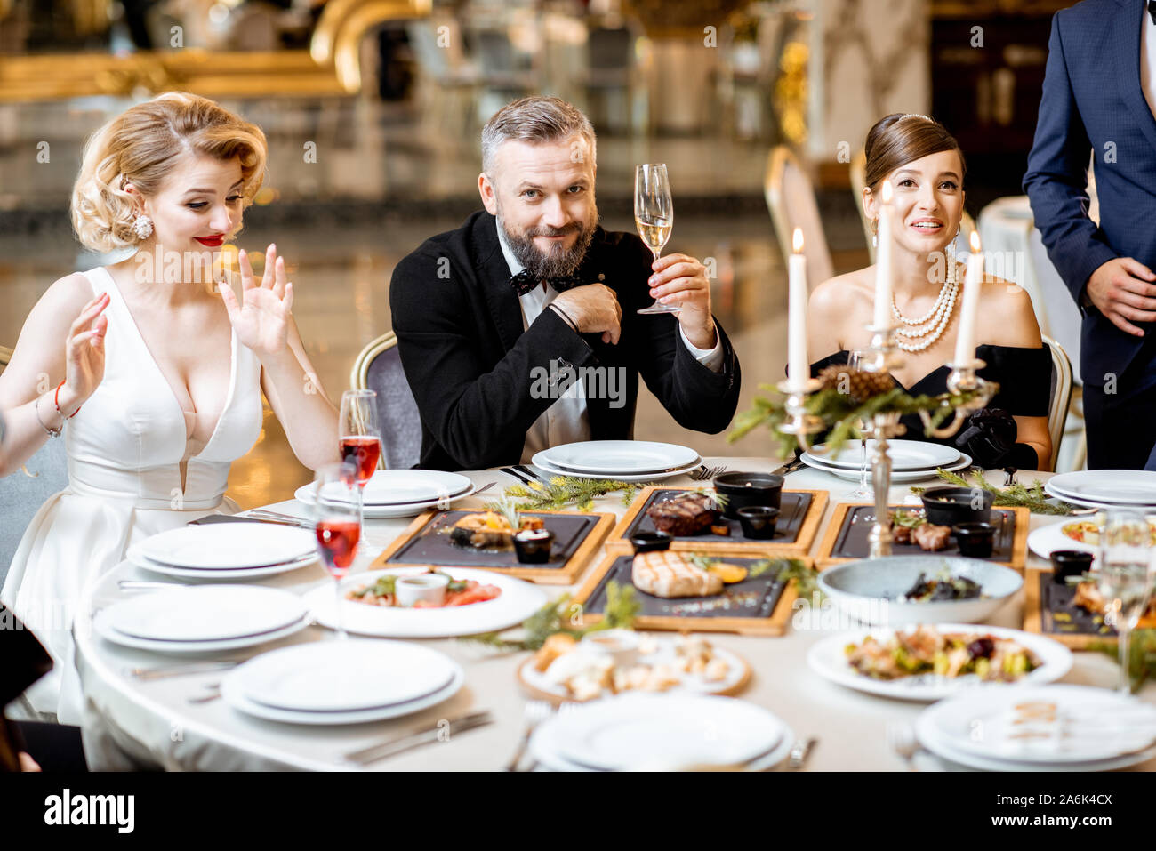 Elegantly dressed group of people having a festive dinner at a well-served table with tasty dishes during New Year Eve at the luxury restaurant Stock Photo