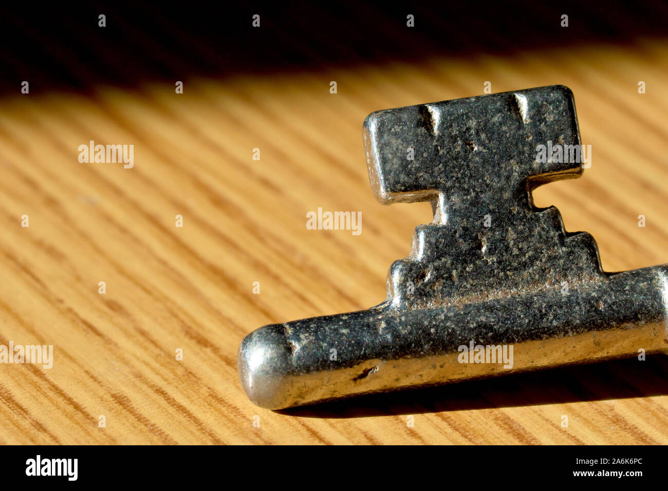 Close up of the bottom end of an old, steel key laying on a brown wooden surface in the sun. Stock Photo