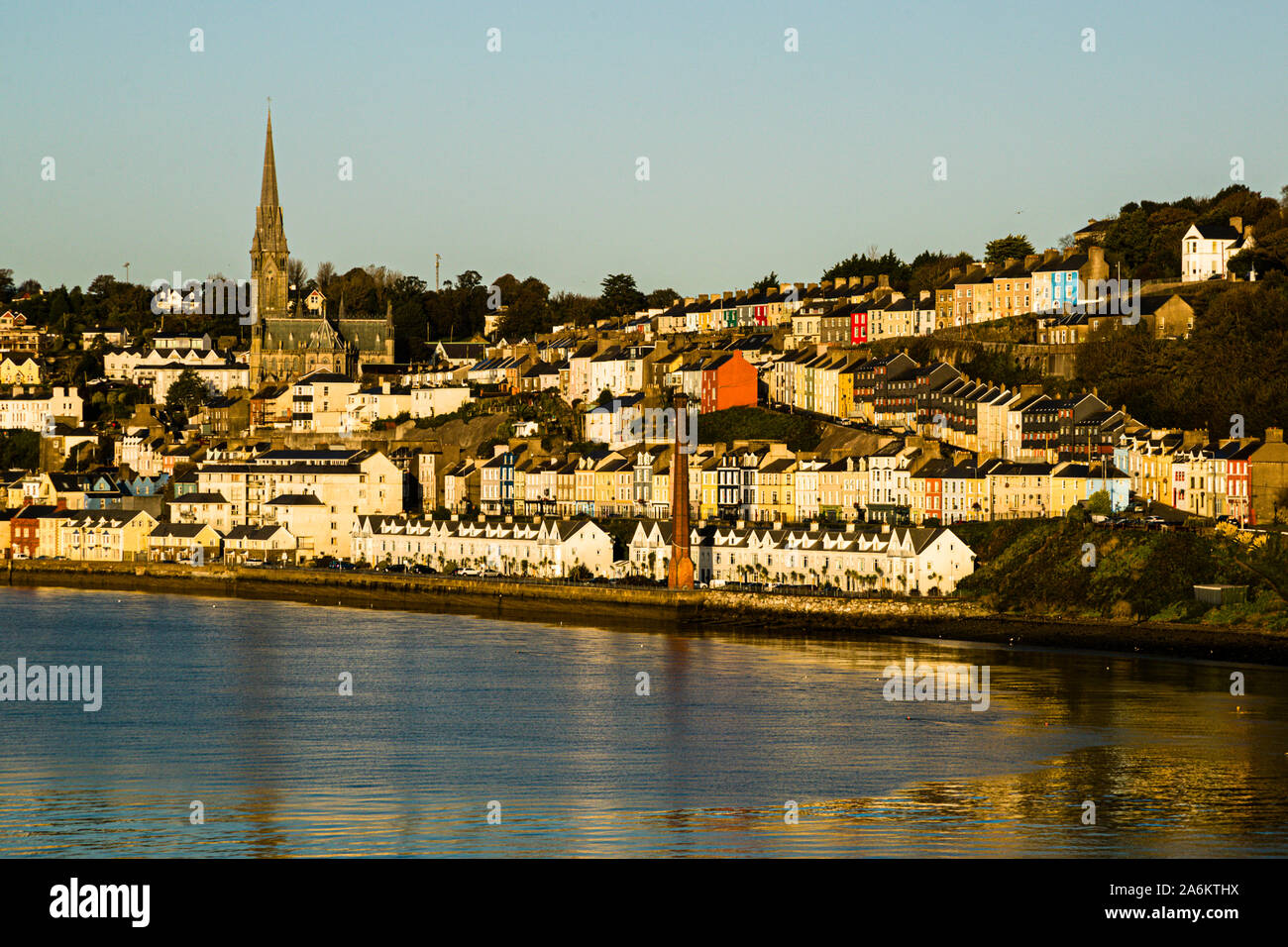 St Colman's Cathedral, its 91-metre spire towering over the colourful terraced houses of Cobh, County Cork, Ireland, seen here with a red factory chimney on the waterfront in the morning light. Built between 1868 and 1919 in the Gothic Revival style (architects Edward Welby Pugin and George Ashlin), the cathedral holds the largest carillon in Ireland and Britain, with 49 bells. Cobh, formerly known as Queenstown, was the Titanic's last port of call in 1912. Stock Photo
