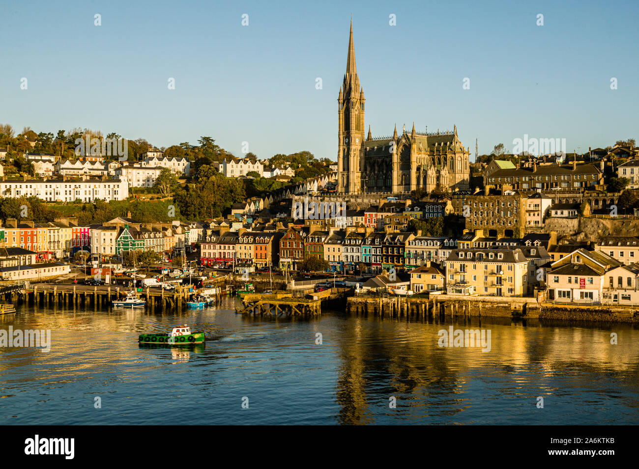 St Colman's Cathedral, its 91-metre spire towering over the colourful terraced houses of Cobh, County Cork, Ireland, known locally as the "Deck of Cards"; a small green boat crosses the water in the foreground. Built between 1868 and 1919 in the Gothic Revival style (architects Edward Welby Pugin and George Ashlin), the cathedral holds the largest carillon in Ireland and Britain, with 49 bells. Cobh, formerly known as Queenstown, was the Titanic's last port of call in 1912. Stock Photo