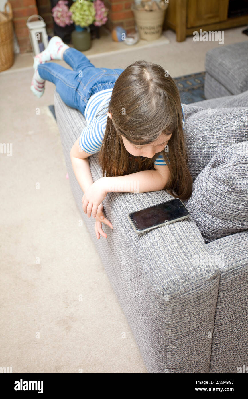 Six year old girl laying on the arm of a sofa watching her phone Stock Photo