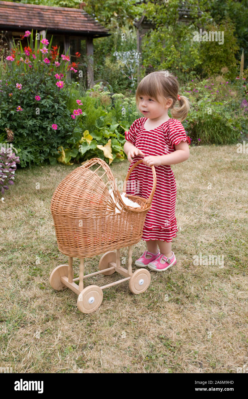 Two year old girl pushing her wooden pram in red dress in garden Stock Photo