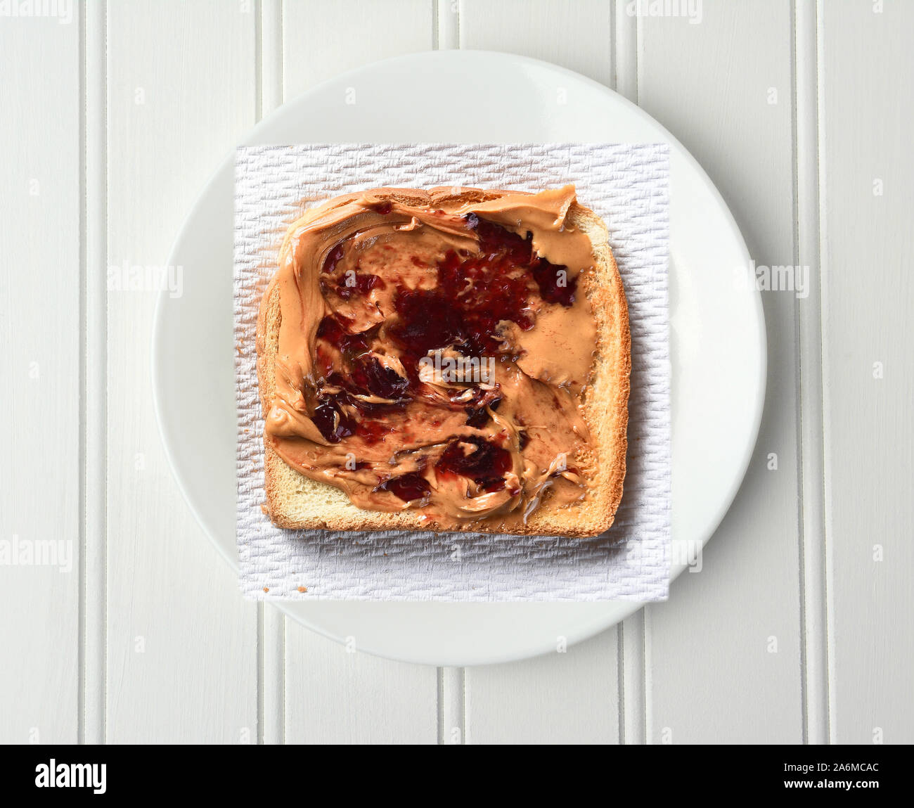 Overhead shot of a slice of toast with Peanut Butter and Jelly, on a paper napkin and white plate. Stock Photo