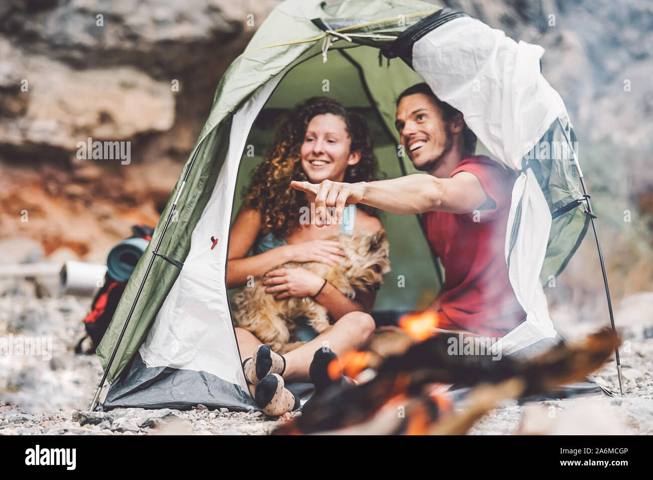 Couple of trekker sitting in the tent with their pet - Happy man and woman having fun in vacation camping around rock mountain next to the fire Stock Photo