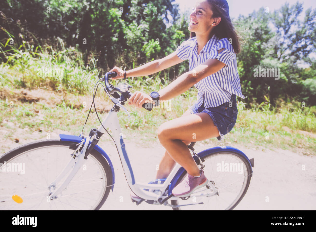Smiling pretty young woman in jeans shorts riding bicycle in the park ...