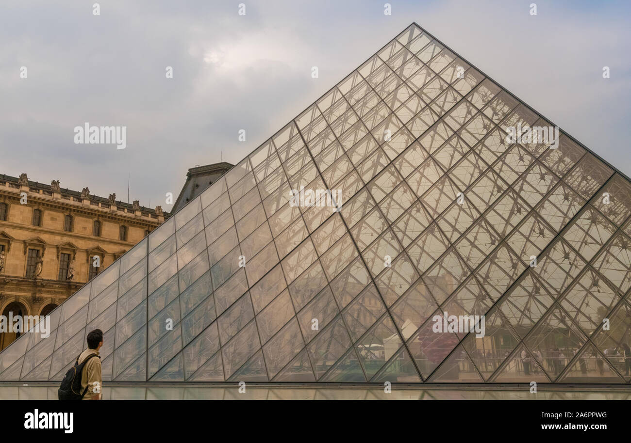 A male visitor with a backpack stands in front of the Louvre Pyramid