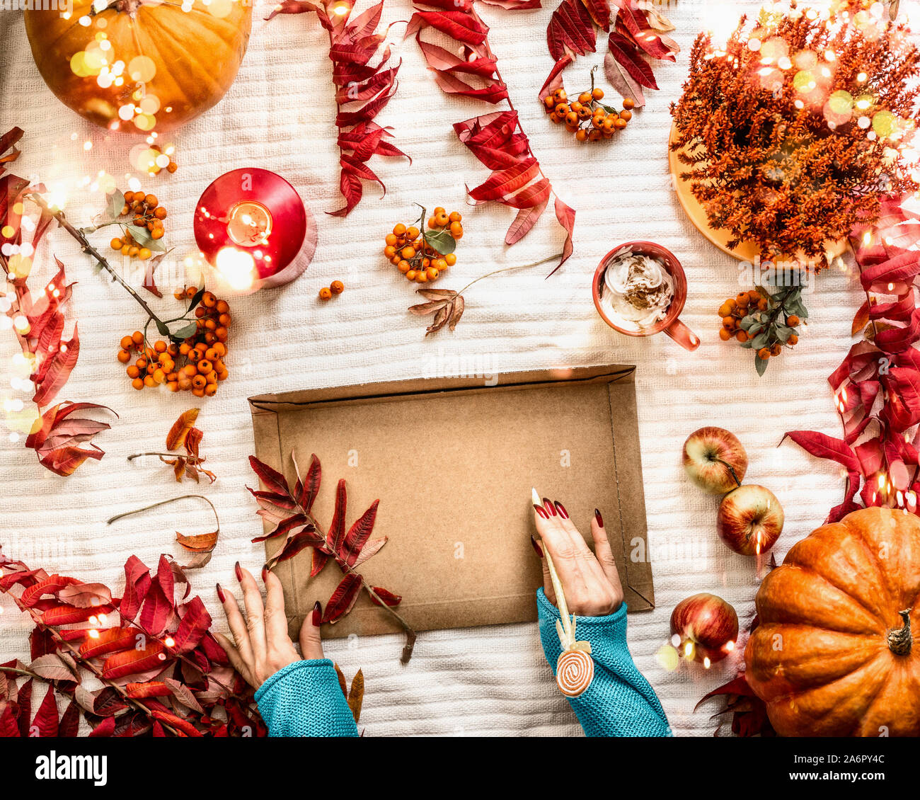 Female hands write at large kraft paper envelope on white desktop with autumn mood arrangement: burning candles, pumpkins, red fall leaves and flowers Stock Photo