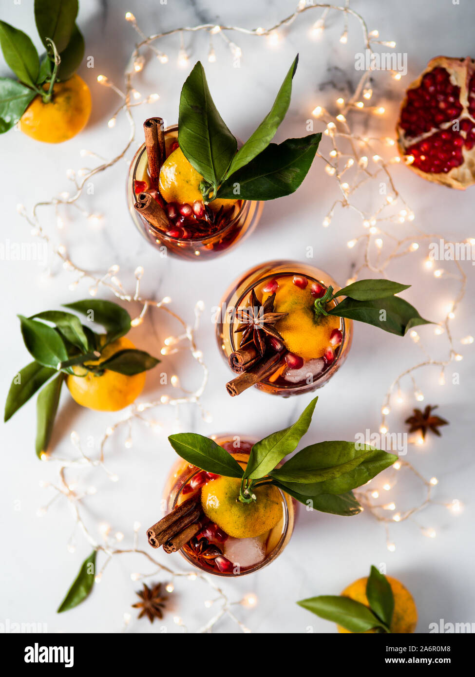 Winter sangria on white marble background. Holiday sangria in glasses with fruit slice, pomegranate and spices on decoration lighting chain tabletop. Top view or flat lay. Vertical. Stock Photo