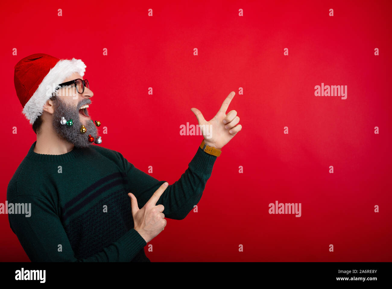 Photo of crazt man screaming and pointing at copyspace over red background and wearing santa claus hat Stock Photo